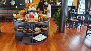 Een ontbijtbuffet met diverse gerechten en dranken, omringd door houten meubels en potplanten.