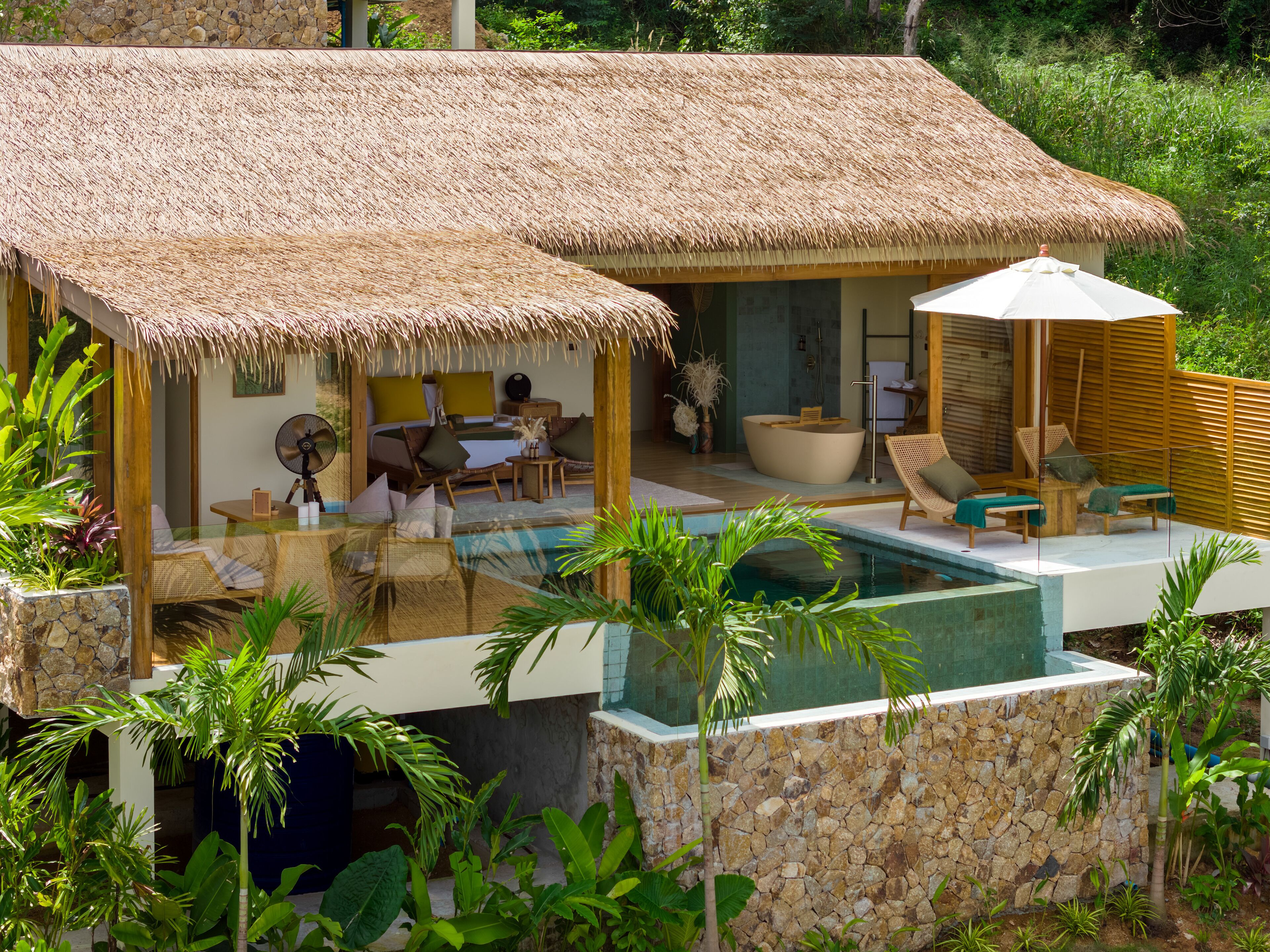 A thatched-roof bungalow with a pool, lounge chairs, and a small table.