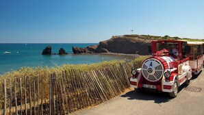 A vintage red and white train parked on a coastal road with a clear blue sky and ocean in the background.