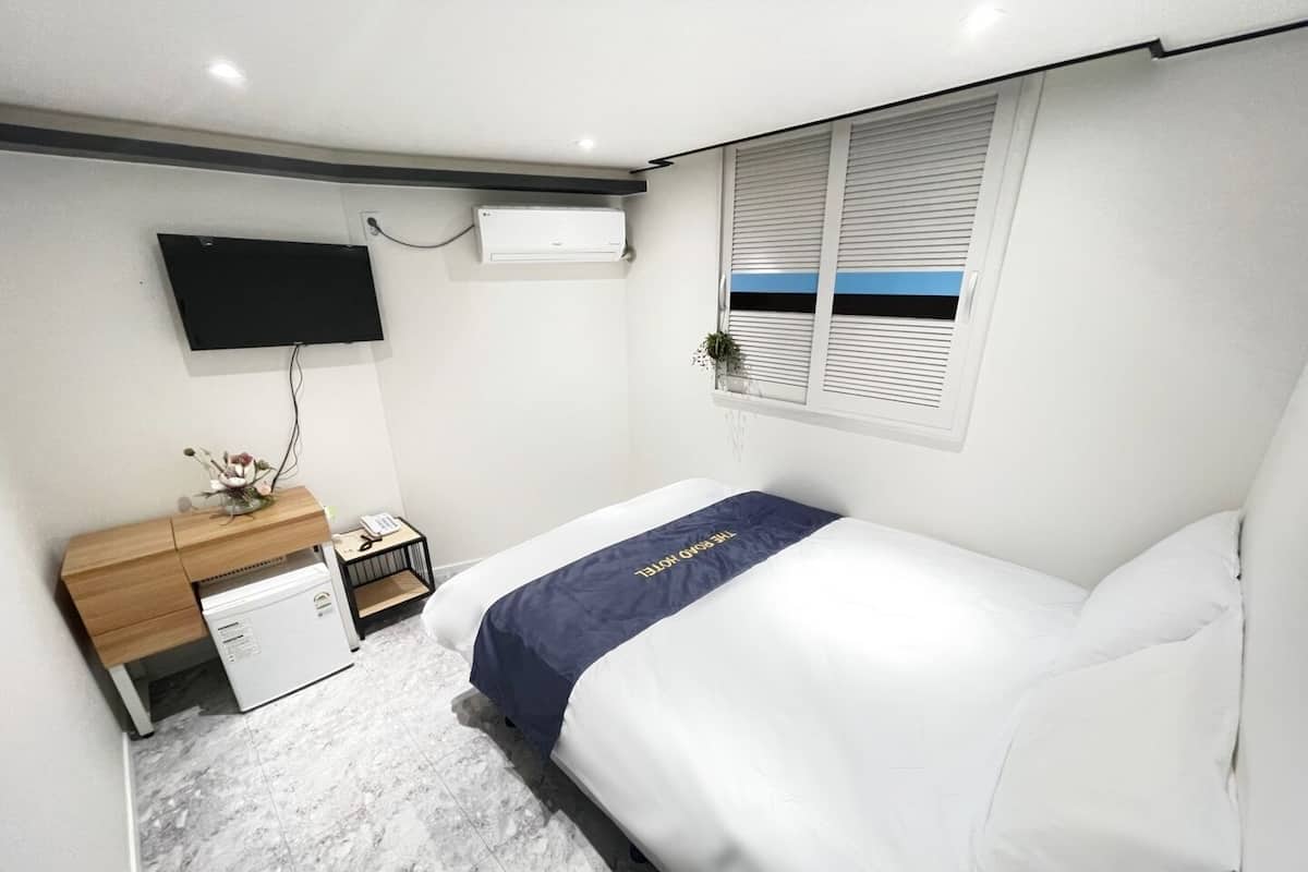 A hotel room with a bed, a desk with a computer, a TV mounted on the wall, and a window with blinds.