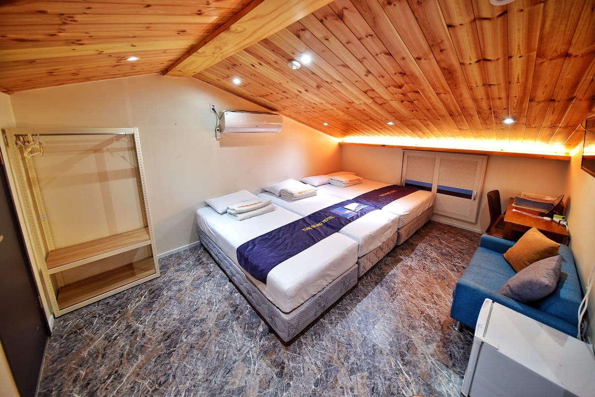 A bedroom with a wooden ceiling, a bed with a blue and white bedspread, a blue sofa with cushions, and a small desk with a chair.