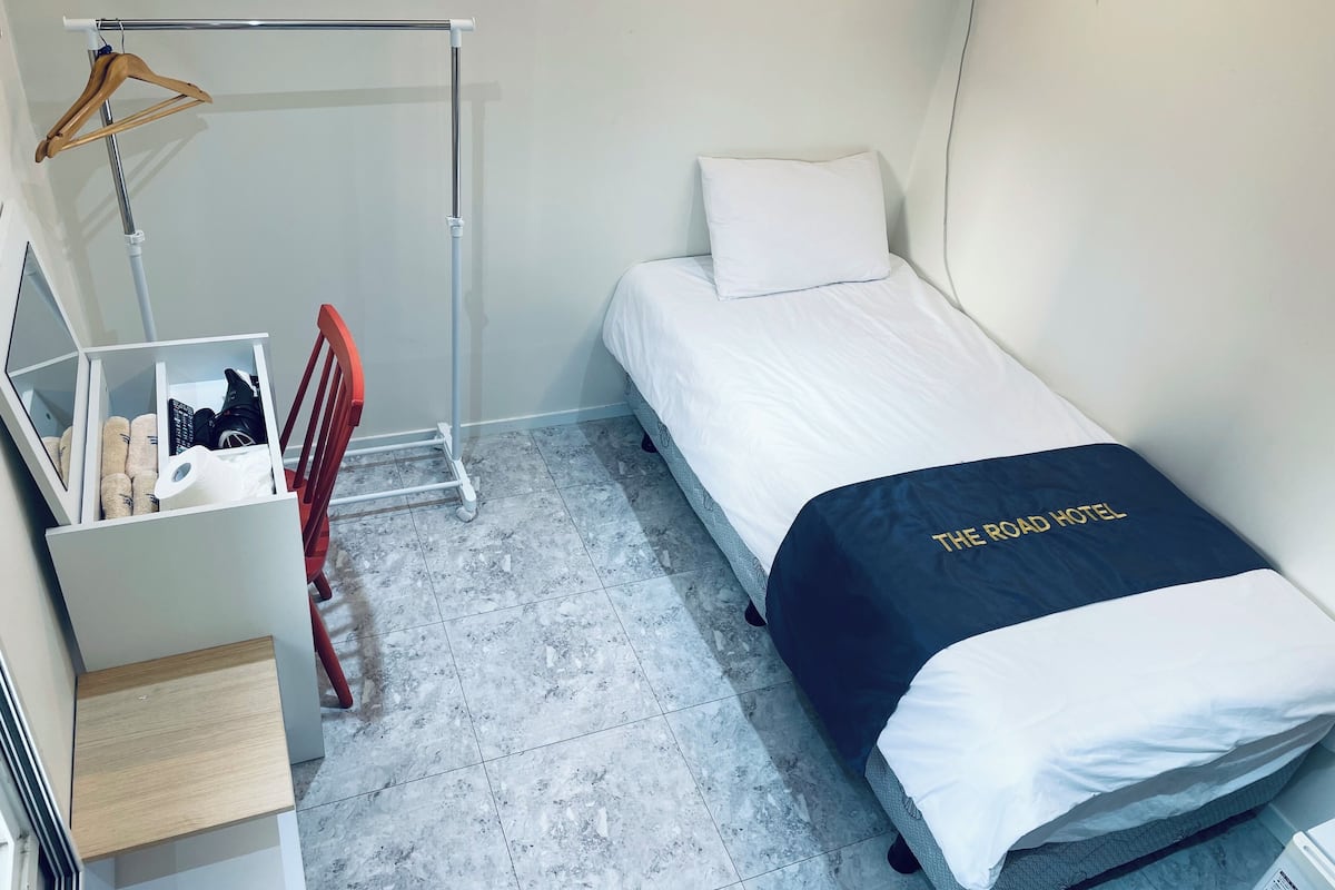A small, neatly organized room with a bed, a wooden hanger on a rack, a small table, and a wardrobe.