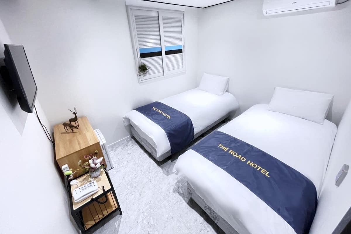 A hotel room with two beds, a desk with a computer, a window with blinds, and an air conditioning unit.
