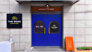 A hotel entrance with a blue door, a welcome sign, and a sign indicating check-in and check-out times. - The Road (Seoul)