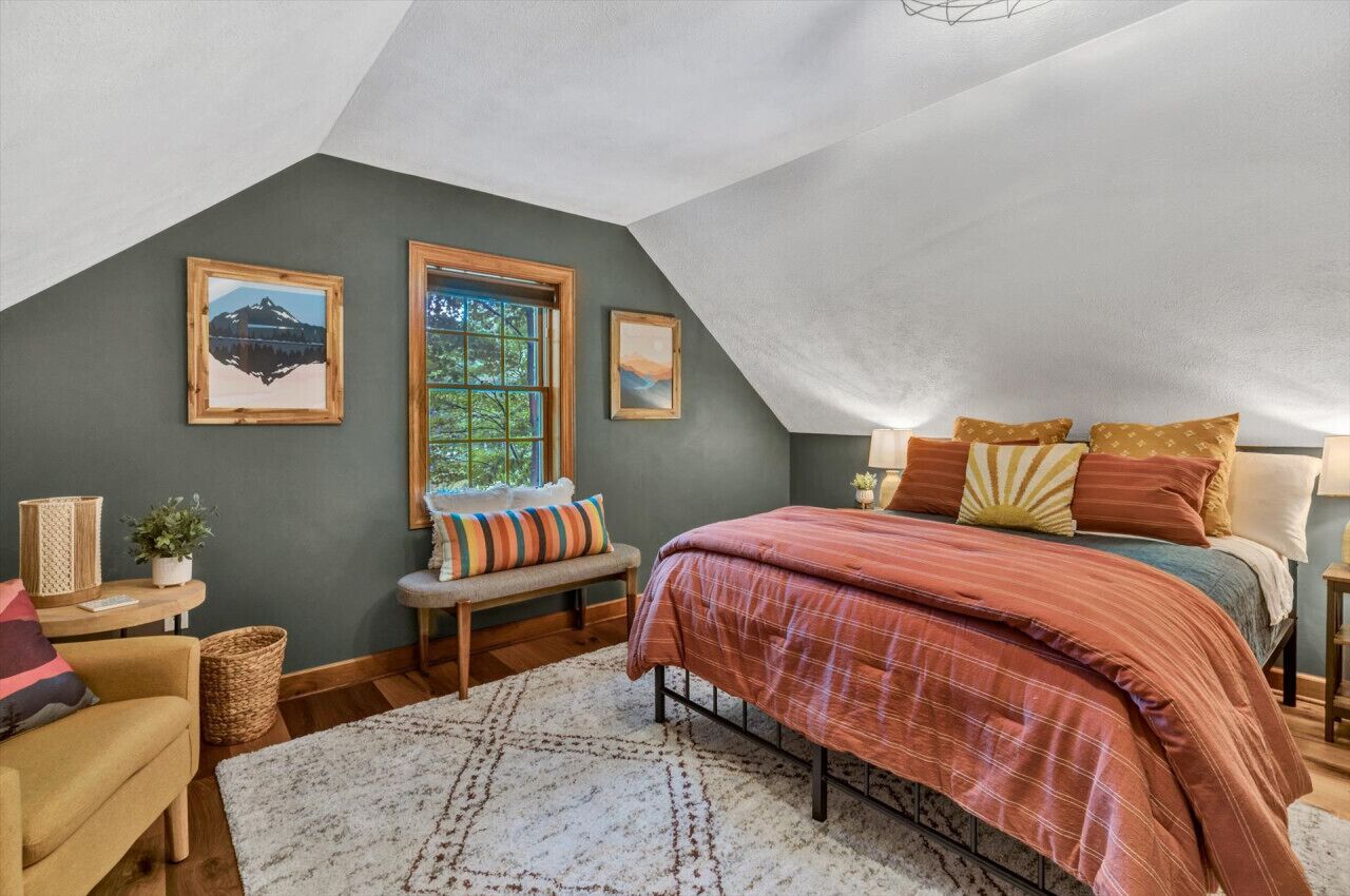 A bedroom with a sloped ceiling, a bed with a plaid comforter, two framed pictures on the wall, a bench with cushions, and a window with a view of greenery.
