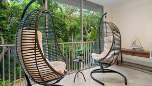 Deux fauteuils suspendus avec des coussins, une petite table avec une plante, et une vue sur la verdure à travers la rambarde d’un balcon.