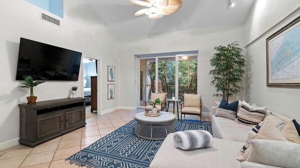A spacious living area with a flat-screen TV, a dark wooden entertainment unit, a round coffee table, a beige sofa, and a large potted plant.