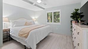 A bedroom with a bed, bedside table, lamp, window with blinds, dresser, and a ceiling fan.