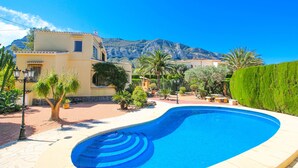 A swimming pool with a curved edge, surrounded by a paved patio with potted plants and a lamp post. In the background, there is a mountain and a building with a yellow facade. - Villa with private pool in Denia (Denia)