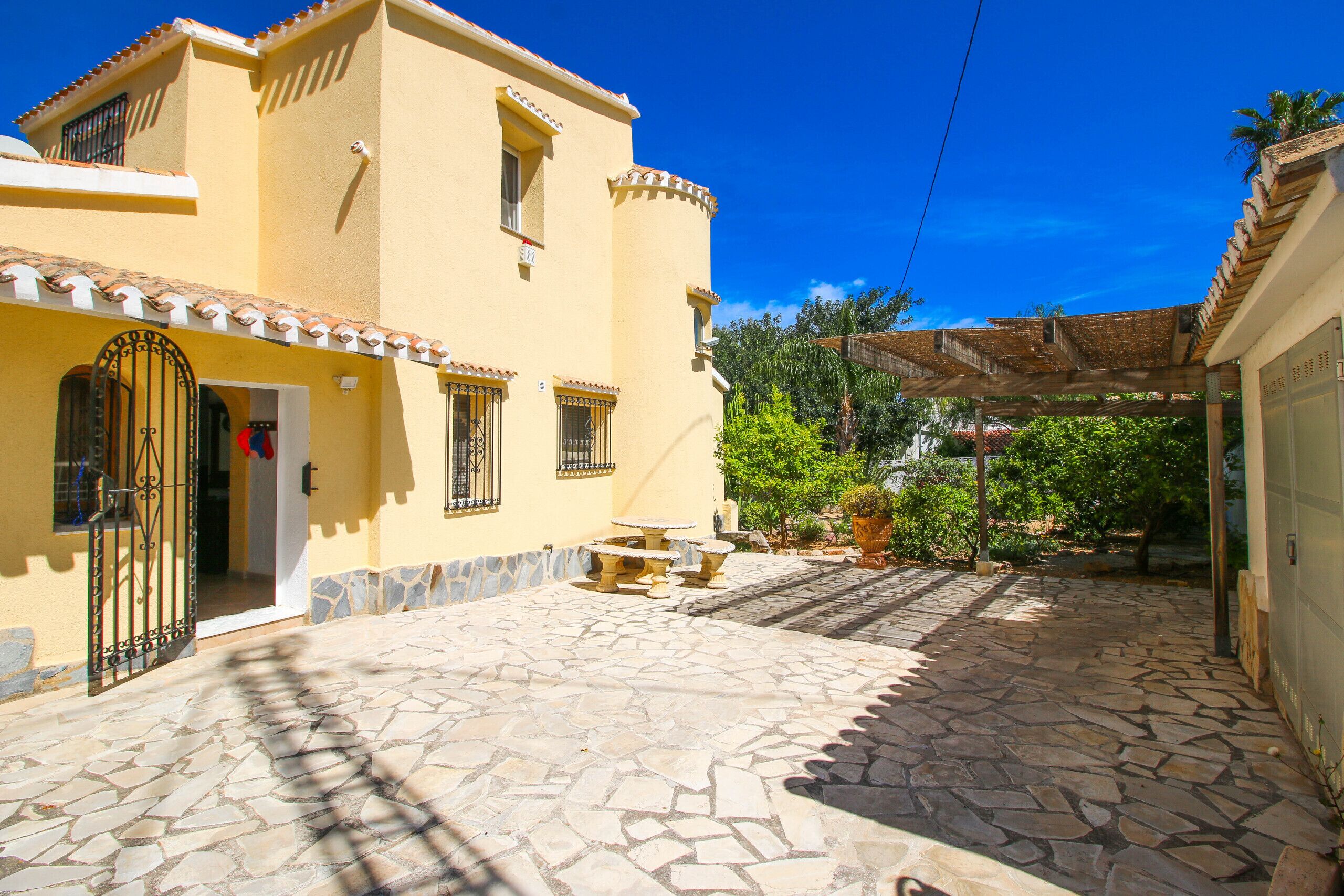 A yellow building with a stone courtyard, a gated entrance, and a covered area with plants.