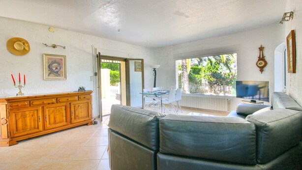 A spacious living room with a leather sofa, a wooden cabinet, and a dining table with chairs. There is a television and a large window providing a view of the outdoors. - Villa with private pool in Denia (Denia)