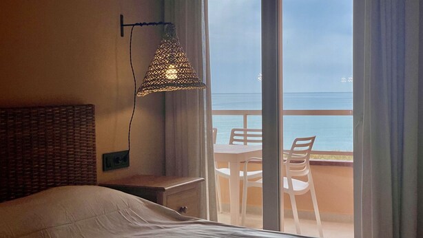 Une chambre avec un lit, un luminaire suspendu, une fenêtre donnant sur la mer et un balcon avec une table et des chaises.