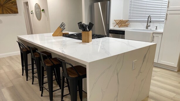 A modern kitchen with a marble island, bar stools, stainless steel appliances, and a window with blinds.