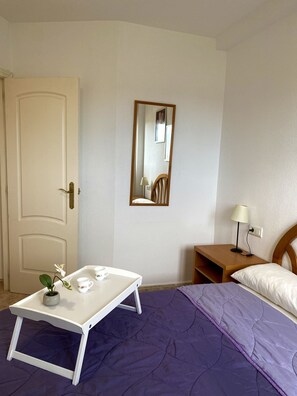 A bedroom with a bed, a bedside table with a lamp, a small table with a plant, and a door.