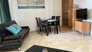 A living room with a sofa, a dining table with chairs, a TV on a wooden cabinet, and a painting on the wall. - MED 19 with paddel court (Denia)