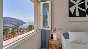 Une chambre avec une grande fenêtre, une table de chevet avec une lampe, un lit avec des oreillers et une œuvre d’art accrochée au mur.