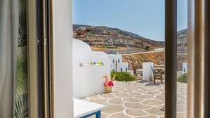 Uitzicht vanuit een kamer met een witte muur en een vloer met patroon, leidend naar een terras met een tafel en stoelen, en een bergachtig landschap op de achtergrond.
