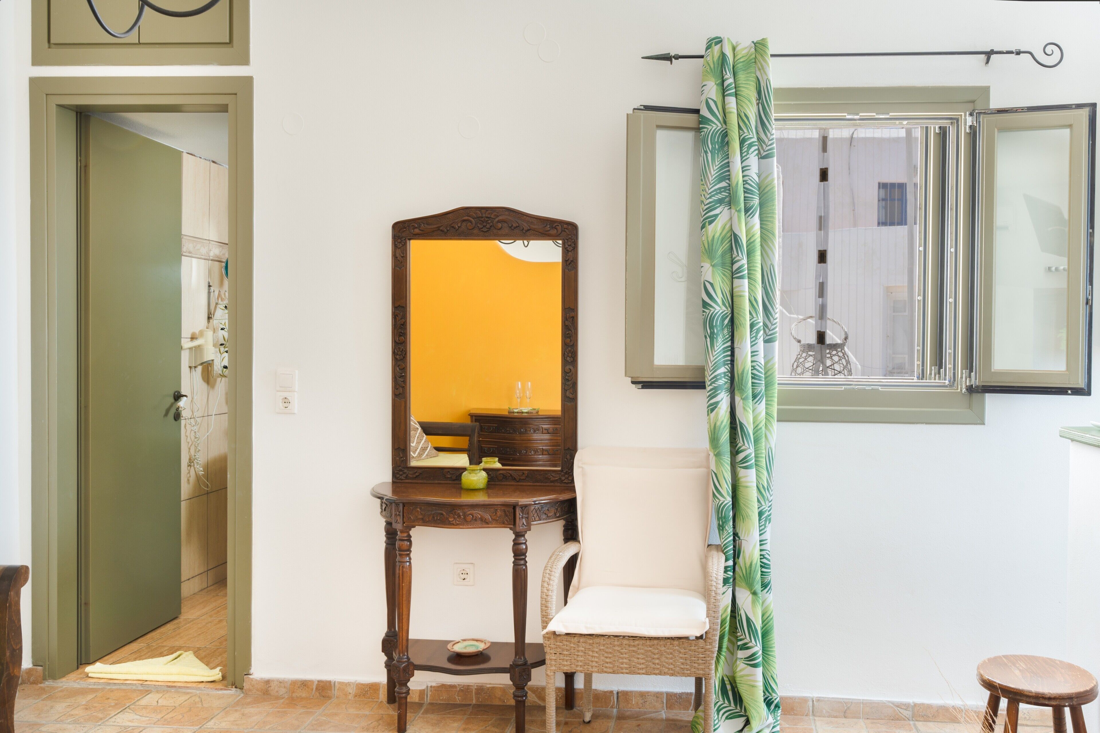 Une pièce comprenant une commode en bois, un miroir, une chaise en osier, un petit tabouret et une fenêtre avec un rideau.