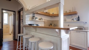A kitchen with a wooden bar counter, white stools, and shelves with kitchenware. - Apartment 'Villa Massa' with Sea View, Wi-Fi and Air Conditioning (Polignano a Mare)