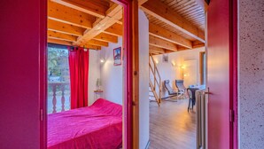 Ein Zimmer mit einem Bett, Holzbalkendecke, einem roten Vorhang, einem Sessel, einem Tisch und einem Heizkörper.