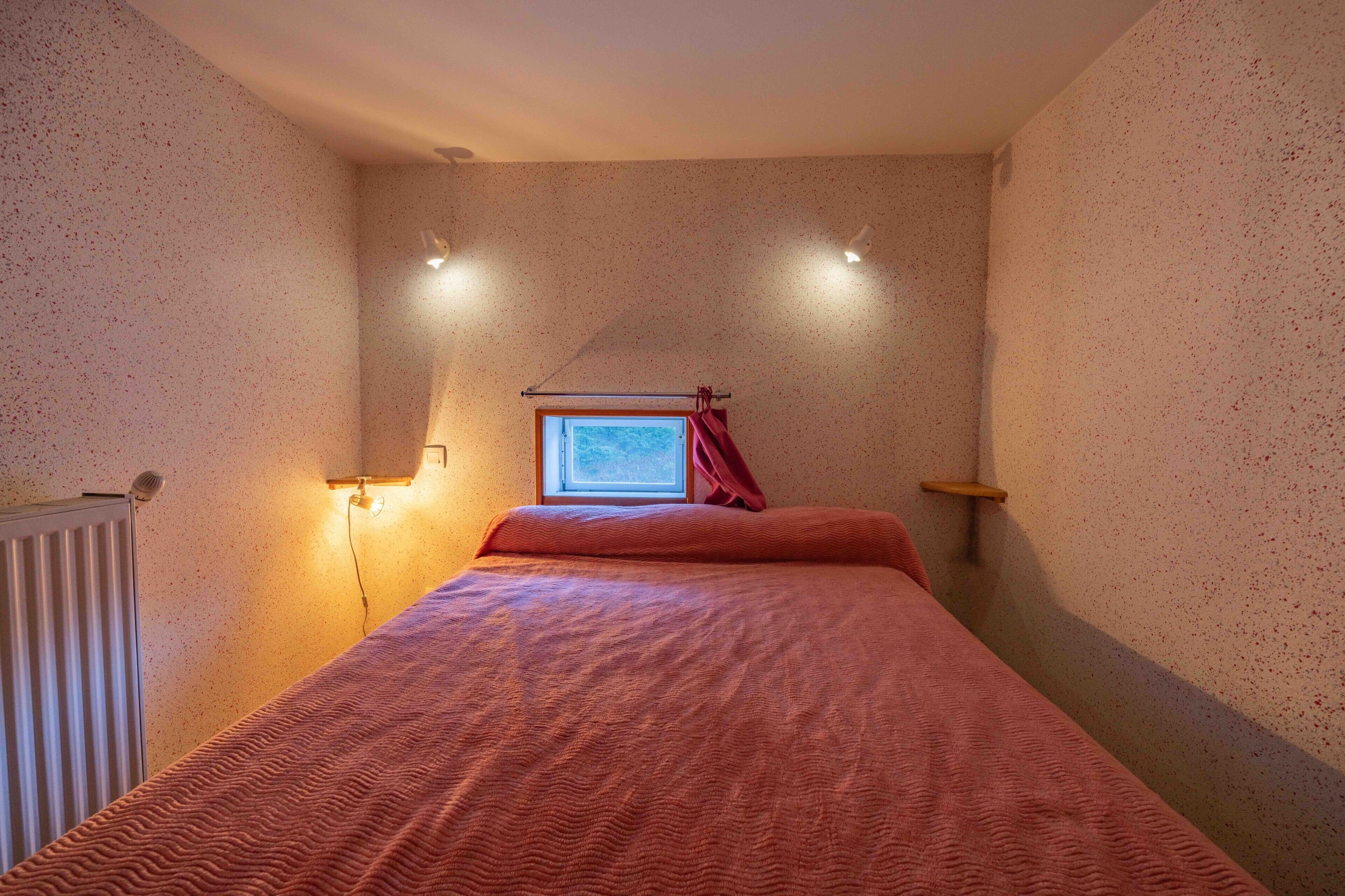 A bedroom with a bed, a window with curtains, a radiator, and wall-mounted lights.
