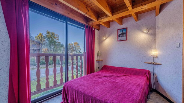 Ein Zimmer mit großem Fenster, einem Bett mit roter Decke, Holzbalken an der Decke und einem kleinen Tisch mit einer Lampe.