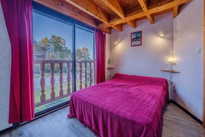 A room with a large window, a bed with a red blanket, wooden beams on the ceiling, and a small table with a lamp.