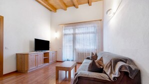 A living room with a wooden floor, a sofa, a TV on a wooden cabinet, and a window with sheer curtains. - Apartment 'Antersi Robbi' with Mountain View, Shared Garden and Wi-Fi (St. Vigil Enneberg)