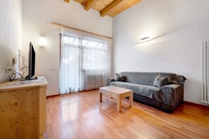Una sala de estar con piso de madera, un sofá gris, una mesa de centro de madera, un televisor sobre un mueble bajo y una ventana con cortinas.
