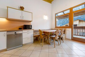 Una cocina con lavavajillas, microondas, mesa de madera con sillas y ventana con vistas a una estructura de madera.