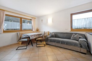 A room with a sofa, a dining table with chairs, a window with curtains, and a cross on the wall.