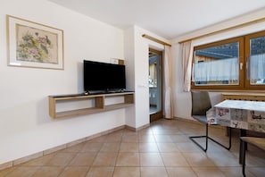 A room with a TV on a wooden shelf, a dining table with a tablecloth, and a window with curtains.