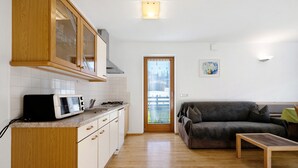 Eine Küche mit weißen Schränken, einer Mikrowelle und einem Herd. Über der Arbeitsfläche befindet sich ein Holzkasten mit Glastüren. Im Wohnbereich stehen ein graues Sofa und ein kleiner Holztisch.