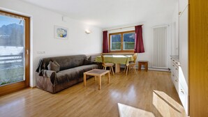 Ein Wohnzimmer mit einer Couch, einem Esstisch, Stühlen, einem kleinen Holztisch, einem Heizkörper und einem Fenster mit Bergblick.