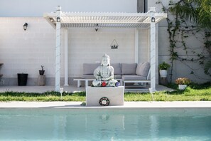 Une statue de Bouddha se trouve dans un belvédère blanc, près d’une piscine, avec un canapé gris et une petite table.