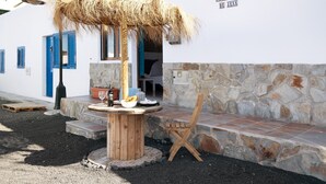 A white building with a stone wall, a wooden table with a glass, a straw umbrella, and a wooden chair. - Holiday Home 'El Rinconcito De Jess' with Wi-Fi and Air Conditioning (El Golfo)
