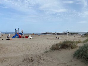 En sandstrand med en legeplads, få mennesker og en bygning i det fjerne.