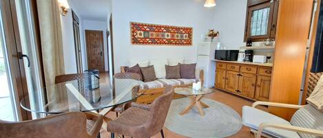 A modern living room with a glass coffee table, brown leather chairs, a sofa with patterned cushions, a kitchen area with wooden cabinets, and a decorative wall hanging.