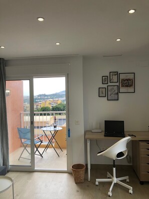 A modern room with a balcony, a desk with a laptop, a chair, and artwork on the wall.