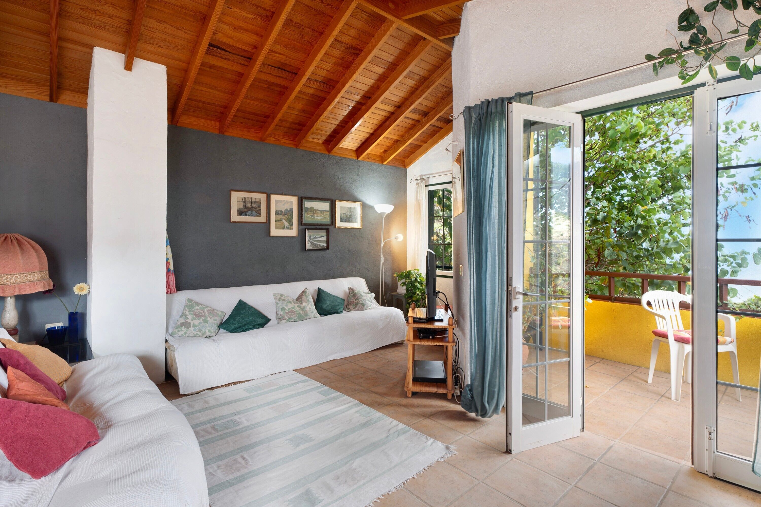Una habitación con techo de madera, un sofá blanco con cojines verdes, un escritorio pequeño con una lámpara y una puerta corredera de vidrio que da a un espacio exterior con una silla.