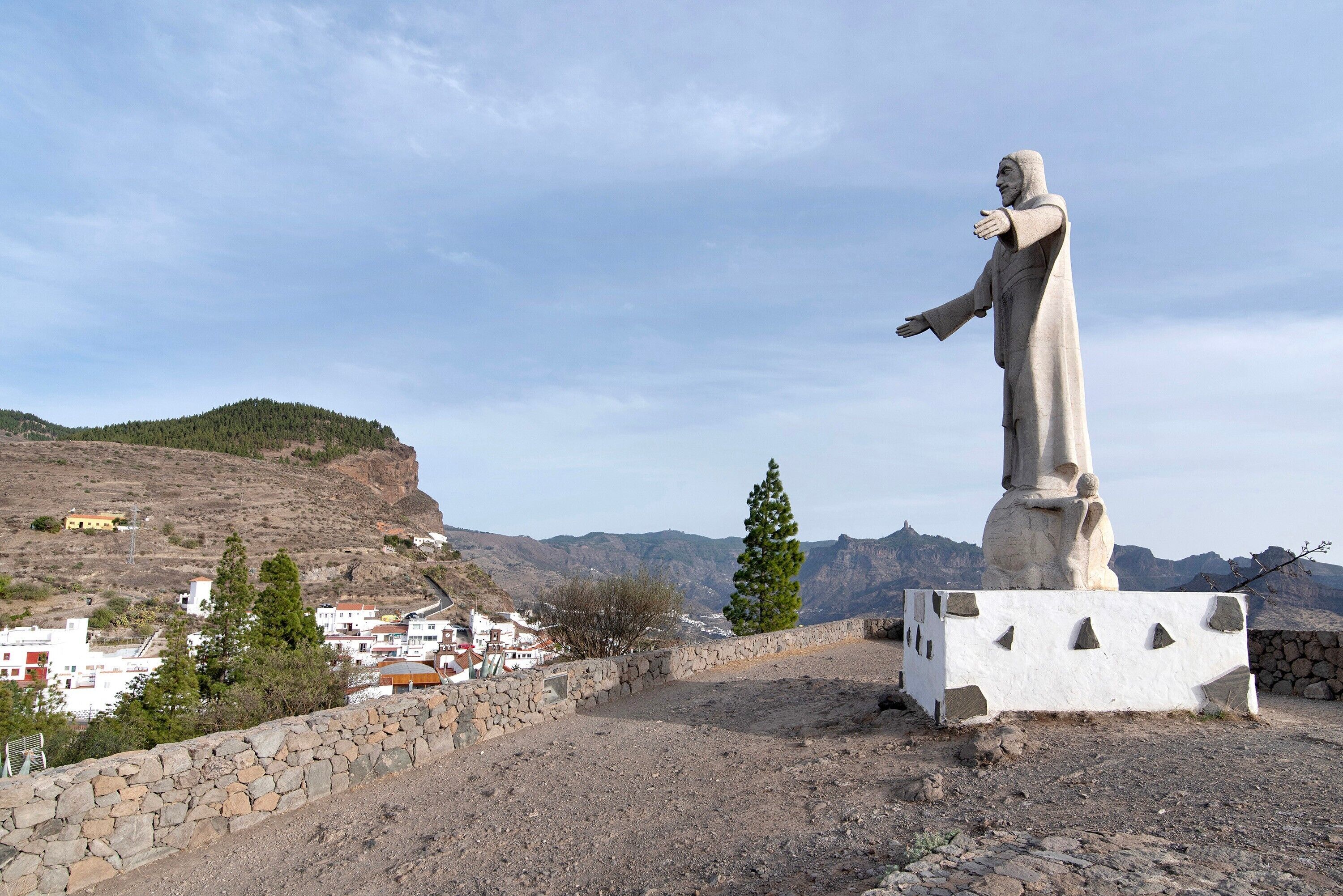 Eine Statue auf einem Sockel, im Vordergrund eine Steinmauer und im Hintergrund ein Dorf.