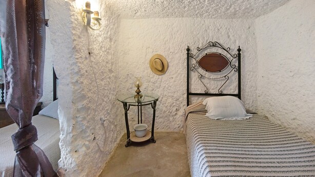 Un dormitorio con cama, una mesita de noche con lámpara, un sombrero colgado en la pared y techo con textura.