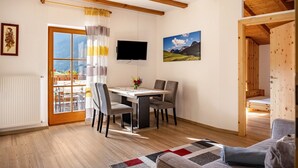 A room with a wooden ceiling, a dining table with chairs, a TV mounted on the wall, and a window with curtains.
