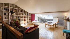 Un salon de style industriel, avec un plafond haut, un banc en bois, un fauteuil blanc, une bibliothèque remplie de livres et une lucarne.