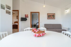Uma área de refeições com uma mesa redonda, cadeiras brancas, um centro de mesa de vime, um sofá cinza e uma TV de tela plana presa na parede.