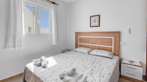 Un dormitorio con cama, mesita de noche, ventana con cortinas, un cuadro en la pared y una pequeña mesa con un libro.
