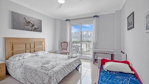 Un dormitorio con una cama grande, una cama más pequeña, una silla, una mesa, un ventilador de techo y una ventana con vistas.