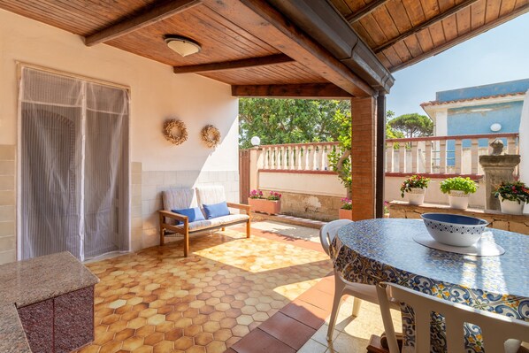 A covered patio with a tiled floor, a wooden ceiling, a table with a blue and white patterned tablecloth, a bench, and a chair.
