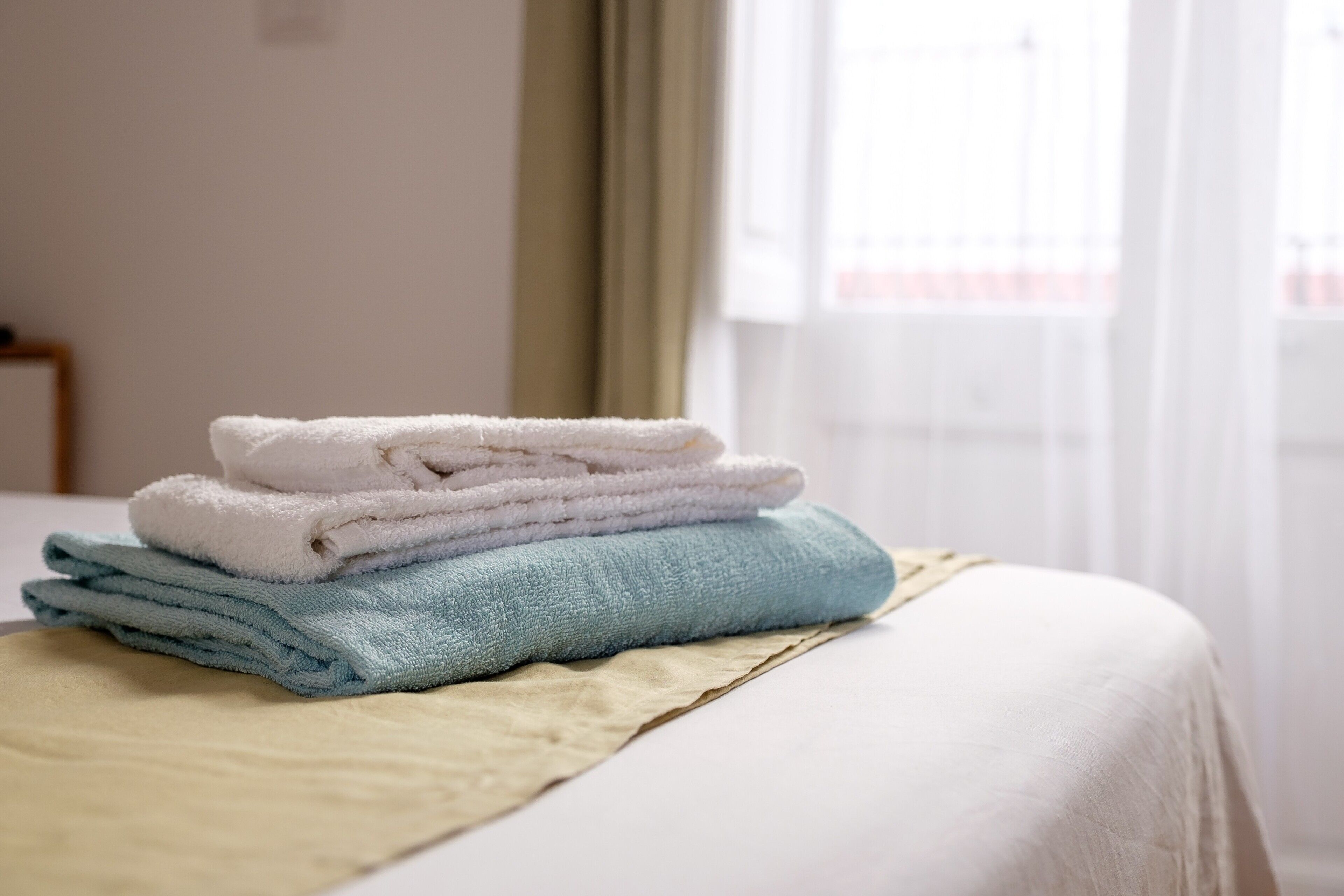 Ein ordentlich bezogenes Bett mit weißer Bettdecke und beiger Decke, ein Stapel gefalteter Handtücher in Weiß, Hellblau und Beige, und ein Fenster mit durchsichtigen Vorhängen im Hintergrund.