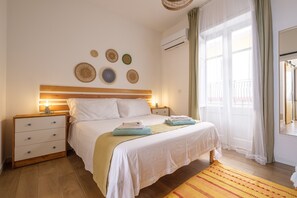 A bedroom with a wooden headboard, a bed with white bedding, a nightstand with drawers, a bedside table with a lamp, and a window with sheer curtains. - Holiday Home 'Mira' accomodation with a terrace. (Spadafora)